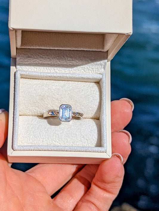 Emerald-Cut Moissanite and Gold Accent Ring  from DEI Collection in an ivory plush jewellery box held above the ocean by Booblinka jewellery