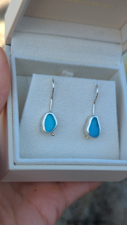 Turquoise Sea Glass Silver Drop Earrings held by hand above the ocean, capturing the vibrant turquoise hues and coastal charm.